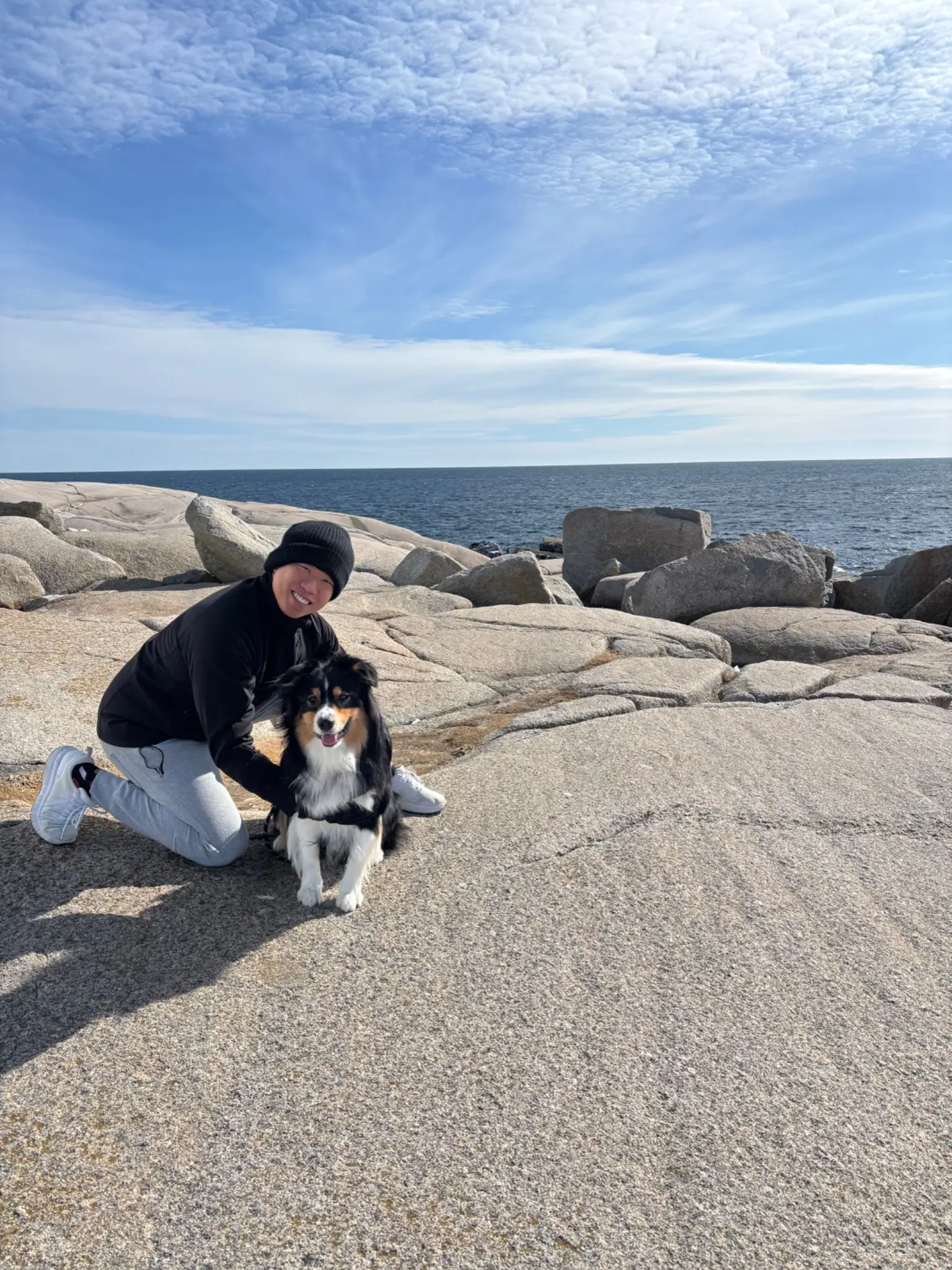Sui Qu kneeling beside Atlas, a tricolor Australian Shepherd, on the granite shore at Peggy's Cove